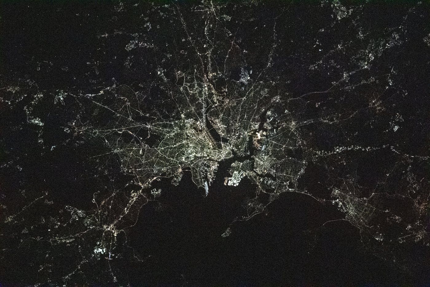 Boston at night from the International Space Station, the lit street grid wrapping around the dark water of Boston Harbor with suburbs spreading inland