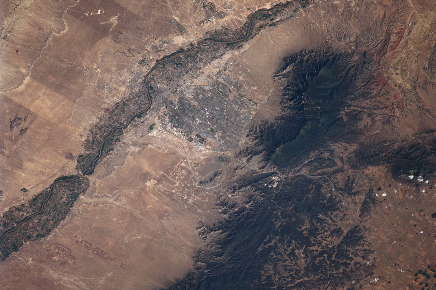 Albuquerque from orbit: the grey grid of the city along the green ribbon of the Rio Grande, with the dark, forested Sandia Mountains rising beside it
