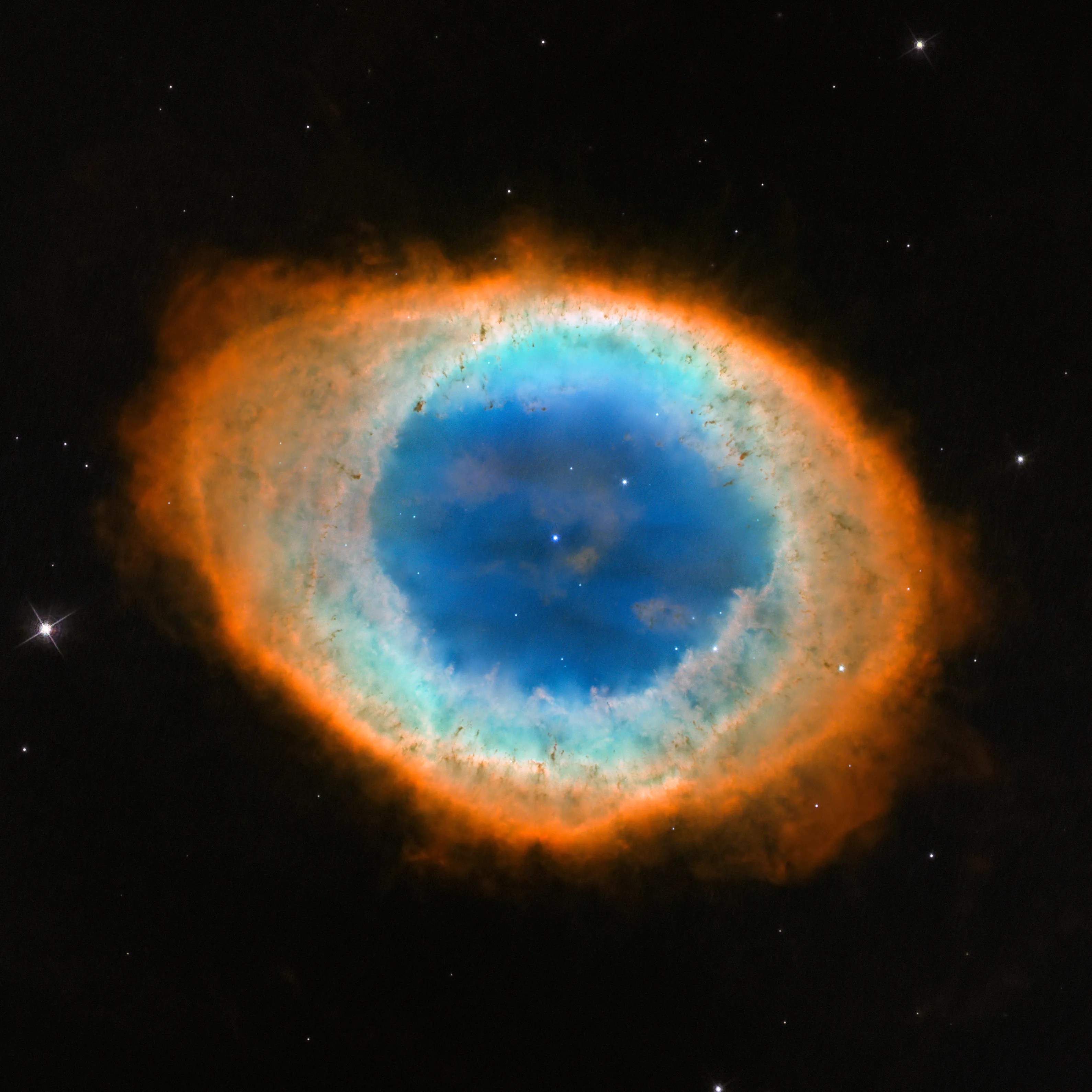 The Ring Nebula M57 as captured by the Hubble Space Telescope — a glowing smoke ring of gas expelled by a dying star, showing blue helium at the centre, green hydrogen and oxygen in the inner ring, and red nitrogen and sulfur in the outer ring