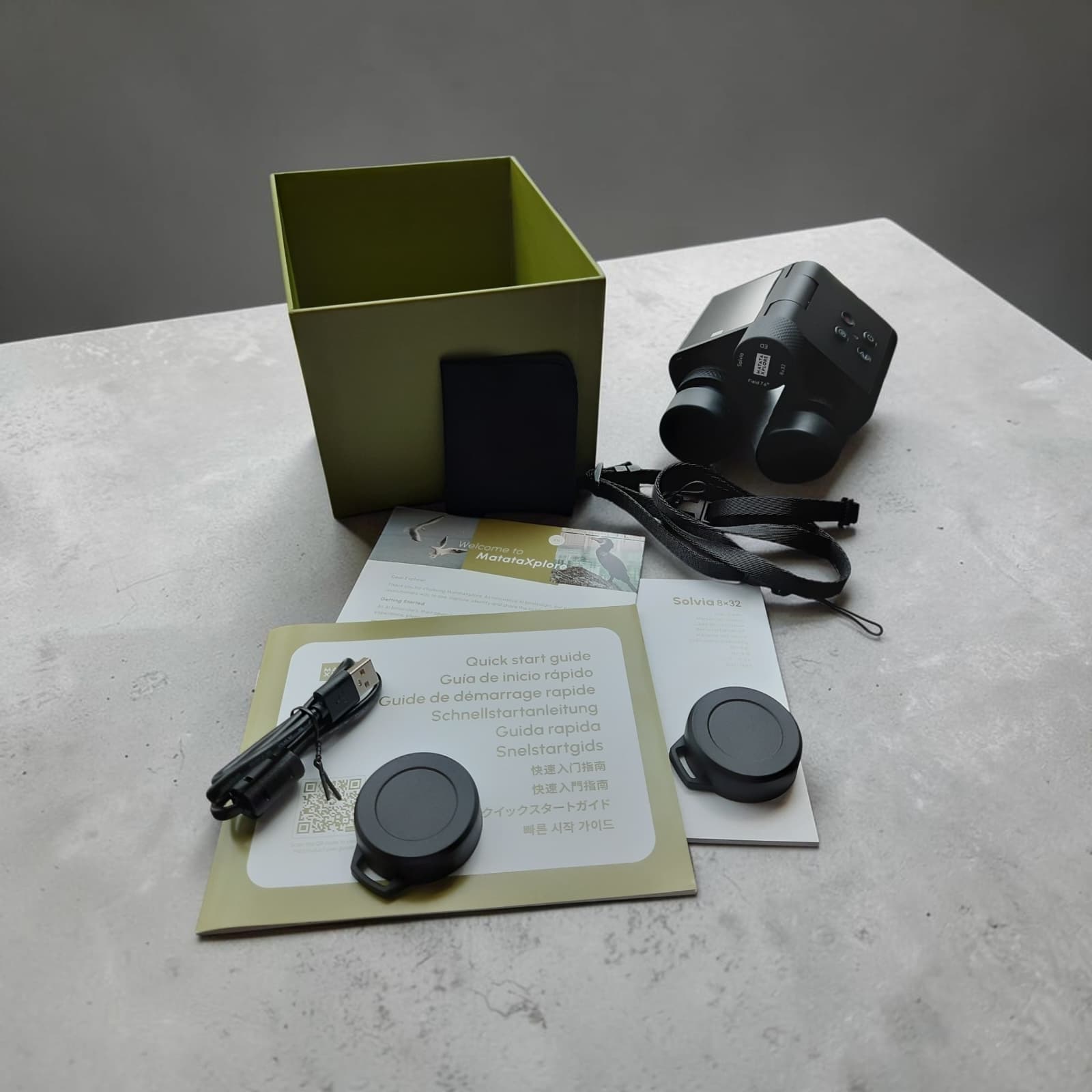 Full unboxed contents of the Solvia 8×32 Base kit laid out: binoculars with neck strap attached, USB-C cable, lens cleaning pouch, quick start guide, welcome card, user manual, and two lens caps